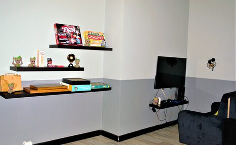Арендовать Лофт-кальянная с игровыми приставками. Игровая комната с серыми стенами, светлым деревянным полом. На стенах расположены черные полки с настольными играми и фигурками. Рядом установлен телевизор на настенном креплении. Слева темный диван. Освещение настенное, стильное.