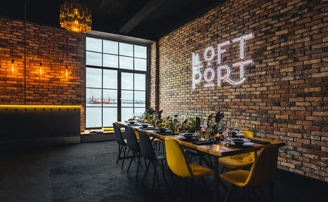 Арендовать Лофт Порт. Loft-пространство с кирпичными стенами, длинным обеденным столом и разноцветными стульями. На стене световая надпись. Большое окно с видом на воду. Подвесные светильники и золотистая люстра. Стены обшиты деревом, пол темный.