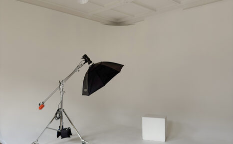 Арендовать Bedford studio (Бедфорд Студия). Светлый зал с белой циклорамой и балконом. Фотостудия с белыми стенами и белым полом. Светло-серое мягкое освещение. В центре студийный осветитель с черным зонтом на стойке. Справа белый куб. Потолок с квадратной панельной отделкой.