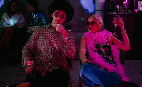 Арендовать Стилизованный лофт в сине-розовых оттенках. Я не могу определить, кто изображён на фото. 

Комната с тусклым освещением, люди сидят на стульях с бокалами; яркая одежда, перья боа, круглые очки. На фоне виден бар или стойка с осветительными приборами. Стиль вечеринки, вероятно, ретро.