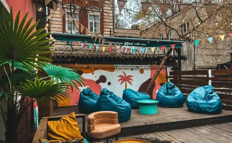 Арендовать Лофт Surf (Серф). Тропический лофт с большой верандой.. Уличная площадка с деревянным настилом, синими креслами-мешками, круглым бирюзовым столом, бежевым креслом и желтыми подушками. Разноцветные флажки над головой, стена с ярким граффити, зелёные растения.