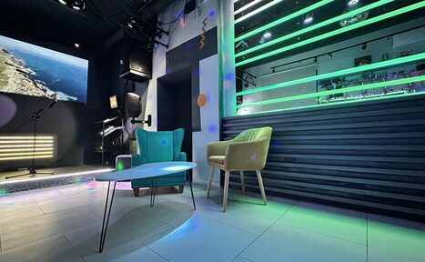 Арендовать Музыкальный лофт на Тульской. Караоке-бар с темным интерьером, сценой и микрофоном. Зеленые световые полосы на стене, бирюзовое и бежевое кресла, белый овальный стол. Стена с изображением природы. Потолок черный, плитка светлая, разноцветные световые эффекты.