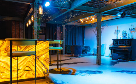 Арендовать Стилизованный двухэтажный лофт. Лофт-пространство с барной стойкой из светящегося камня, высокими стульями, черным пианино, креслами, светодиодным освещением на потолке, декор из веток, открытые балки, пол из светлого материала с темными полосами, приглушенное освещение, свечи.