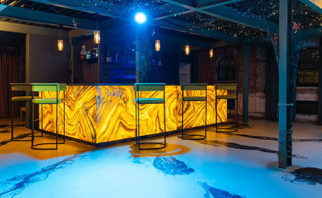 Арендовать Стилизованный двухэтажный лофт. Барная стойка с подсветкой янтарного цвета, высокие стулья, бетонный пол с синими прожекторами, подвесные светильники, открытые стеллажи с бутылками, декорированное помещение с металлическими конструкциями и светодиодными гирляндами.