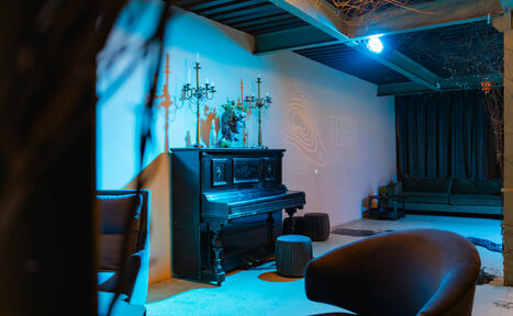 Арендовать Стилизованный двухэтажный лофт. Кабинет с фортепиано у стены, два кресла, пуфы, диван, подсвечники, декор на стене с абстрактным узором и надписью. Мягкое освещение с синим оттенком, бетонный пол, шторы на окне, видна часть лестницы и колонны.