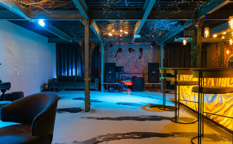 Арендовать Стилизованный двухэтажный лофт. Лофт-пространство с барной стойкой, высокими стульями, мягкими креслами, диваном, колонками и пианино. Потолок с декоративными элементами и гирляндами. Стены белые, пол светлый с темными узорами. Освещение теплое и холодное. На стене проекция.