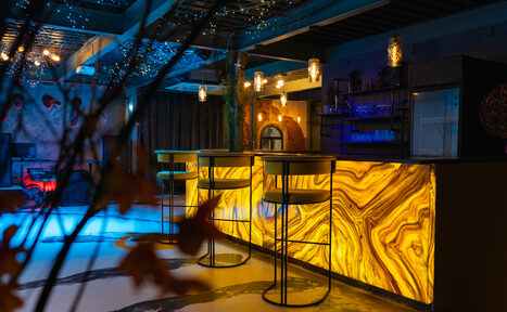 Арендовать Стилизованный двухэтажный лофт. Барное пространство с подсвеченной стойкой в мраморном стиле и высокими стульями. Принт из листьев на переднем плане. Разноцветная подсветка, висячие светильники. Пол — светлый, бетонный. Потолок с декоративными гирляндами. Стойка с полками и микроволновкой.