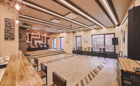 Арендовать Loft Lodge 6. Это просторное помещение с деревянным полом и ступенчатой планировкой. Имеется барная стойка с высокими стульями, сцена, столы и стулья. Потолок украшен балками и современными светильниками. Стены светлые, декорированы картинами. Надписи на меню слева от сцены.