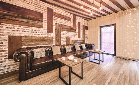 Арендовать Loft Lodge 6. Гостиная с кирпичными стенами и деревянными балками на потолке. Большой коричневый кожаный диван с подушками вдоль стены. Два деревянных стола перед диваном. Подвесное освещение с лампочками в индустриальном стиле. Пол тёмного дерева, входная дверь.