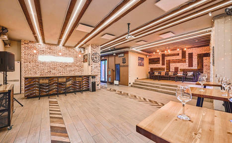 Арендовать Loft Lodge 6. На фотографии изображено просторное помещение с кирпичными стенами и деревянным полом. Видны деревянные столы и стулья, барная стойка с высокими стульями. Потолок украшен светильниками и деревянными балками. Чистое и аккуратное состояние.