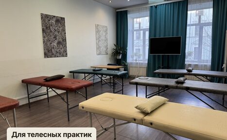 Арендовать Зал для мероприятий, тренингов, семинаров. Просторная комната с несколькими массажными столами разных цветов, расположенными по периметру. Светлые стены с декоративными панелями, большие окна с зелеными шторами, потолочные светильники. На полу — коричневый ламинат. Телевизор на стойке.