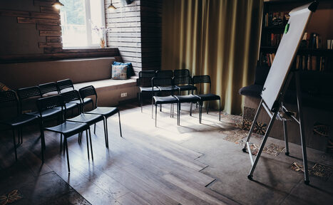 Арендовать Лофт с возможностью зонирования. Небольшое помещение с мягкими скамьями вдоль стены, ряд черных стульев, переносная доска на треноге, деревянный пол, книга на полке, окно с дневным светом, зелёные шторы. Сочетание тёмных и светлых цветов.