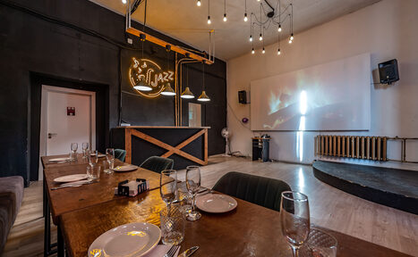 Арендовать Rubin Loft (Рубин Лофт). Зал Джаз. Пространство кафе с деревянными столами, сервированными посудой и бокалами. На стене проекция изображения, рядом с ней аудиоколонка. Черные стены, декоративное освещение с лампами накаливания. Потолок светлый, пол деревянный. Барная стойка.