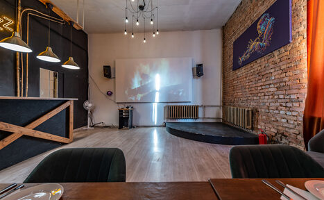 Арендовать Rubin Loft (Рубин Лофт). Зал Джаз. Пространство для мероприятий с барной стойкой и мягкими стульями. На стене кирпичная кладка и белая панель с проектором. Подвесные лампы над баром, люстра с лампами на потолке. Деревянные и металлические элементы, оранжевые шторы.