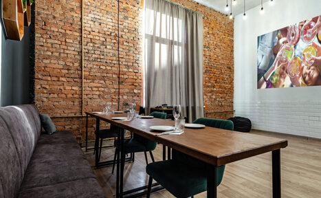 Арендовать Rubin Loft (Рубин Лофт). Лофт средний. Лофт-пространство с кирпичными стенами и деревянным полом. Длинный стол с тарелками и бокалами, по обеим сторонам - стулья и диван. Большое окно со шторами. На стене фото бокалов. Потолочные светильники на проводах.