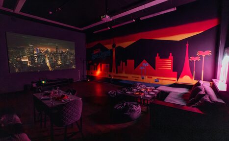 Арендовать Зал Лас-вегас. Лофт-зал в американском стиле . Темная комната с проектором, показывающим изображение мегаполиса. На стене красочный настенный рисунок в виде городского пейзажа. Мебель: столы и стулья, диван, подушки. Тусклое освещение, теплые тона.