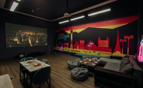 Арендовать Зал Лас-вегас. Лофт-зал в американском стиле . Комната с темными стенами и деревянным полом. На стене яркий городской пейзаж. Большой экран с изображением города. Обеденный стол со стульями, диван, мягкие кресла и обилие подушек. Подвесные светильники.