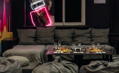 Арендовать Rubin Loft (Рубин Лофт). Зал Лас-вегас. Современное пространство с серыми диванами и пуфами, низким столом с закусками и напитками. На стене неоновая женская фигура и яркий арт. Тёмные стены, окно с белой рамой. Интимное освещение.