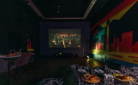 Арендовать Rubin Loft (Рубин Лофт). Зал Лас-вегас. Просторная затемненная комната с проектором, показывающим городской пейзаж. На стене граффити с силуэтами зданий. Расставлены столы, сервированные блюдами и стеклянной посудой. Стены темные, пол деревянный, освещение приглушенное.