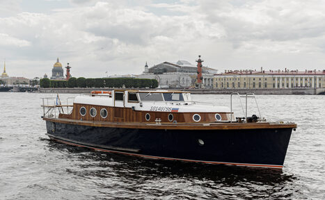 Арендовать Адмиральский катер. Лодка на воде с деревянной палубой и черным корпусом. На фоне городские здания и колонны. Облачно. У лодки круговые окна и открытая верхняя часть с ограждением. На борту текст с цифрами.