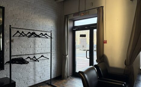 Арендовать Лофт Повод Есть. Стойка с вешалками у белой кирпичной стены. Двойные кожаные кресла справа. Темный деревянный пол. Дверь со стеклянной вставкой, закрытая шторами, надписи "Вход" и "Выход" над дверью. Помещение освещено естественным светом.