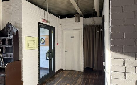 Арендовать Лофт Повод Есть. Коридор с кирпичными стенами, накрытыми белой краской. Потолок с открытыми балками. Деревянный пол темного цвета. Черная стеклянная дверь, белая деревянная дверь, коричневая занавеска. Светлая полка слева. План-схема на стене.