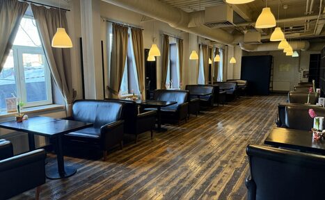 Арендовать Лофт Повод Есть. Пространство в стиле кафе с черными кожаными диванами и деревянными столами, расположенными вдоль окна. Большие окна с бежевыми шторами. Потолочные светильники с теплым светом. Полы из темного дерева. Стены светлые. На столах вазы с цветами.