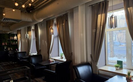 Арендовать Лофт Повод Есть. Пространство кафе с рядами столов и черными креслами вдоль окон. Интерьер с темными шторами и подвесными светильниками. Потолок с видимыми трубами и лампами. Пол темного древесного оттенка, стены светлые. На подоконниках и столах декор.