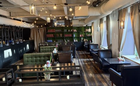 Арендовать Лофт Повод Есть. Пространство с деревянным полом, стеллажами с книгами и темной мебелью. Зеленые мягкие диваны, кожаные кресла и деревянные столы. Лампы и светильники на потолке, большие окна с шторами. Декор: свечи и цветы в вазах. Окрашенные в белый потолочные трубы.