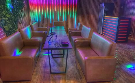 Арендовать Forest(Лес). Современное помещение с диванами из коричневой кожи по обе стороны длинного стола. Вертикальные деревянные панели на стенах. Яркие светодиодные светильники разноцветно освещают пространство. На столе кальян и стеклянные стаканы. Напольное покрытие из дерева.