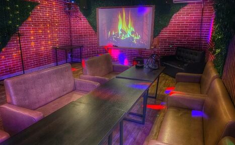 Арендовать Forest(Лес). Караоке-зал с кирпичными стенами, диванами, столом и проектором с изображением камина. На полу деревянное покрытие, освещение разноцветное, стены украшены зеленью.