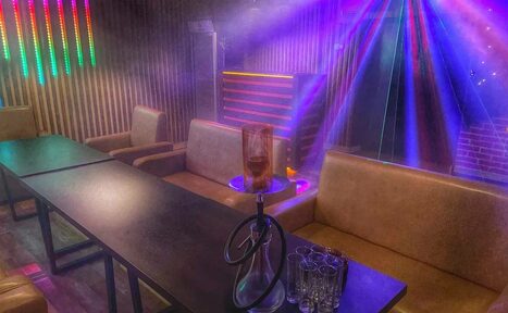 Арендовать Forest(Лес). Тёмное помещение с диванами и столами, на столе кальян и стаканы. Вертикальные деревянные панели на стене, яркие разноцветные светодиодные лампы, фиолетовые световые прожекторы. Пол из тёмного дерева, кирпичная стена.