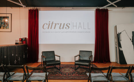 Арендовать Citrus Hall (Цитрус Зал). Зал Grapefruit (Грейпфрут). Пространство с деревянным полом, двумя черными креслами и несколькими складными стульями. Красные шторы по бокам экрана с надписью. Черный барный уголок и ковер с узором. Настенное освещение и серые потолочные балки.