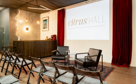 Арендовать Citrus Hall (Цитрус Холл). Зал Грейпфрут. Пространство для презентаций с проектором и экраном. Зал с деревянными стульями, двумя креслами, барной стойкой. Декор: красные шторы, ковёр, висячие светильники. Стены серые и белые, пол деревянный. На экране текст.