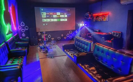Арендовать Rave(Рейв). Караоке-зал в стиле лаундж с кожаными диванами, низким столом и кальяном. Проектор на стене с интерфейсом меню, разноцветная неоновая подсветка, настенные рисунки с картами, черные стены и полы, фоновое освещение, кондиционер вверху.