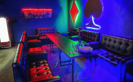 Арендовать Rave(Рейв). Караоке-зал в тёмных тонах с мягкими кожаными диванами и длинным столом. Неоновая подсветка в виде пики и ромба на стенах. На столе бокалы и тарелки. Холодильник в углу, неоновая вывеска с постерами выше. Каменный или бетонный пол.