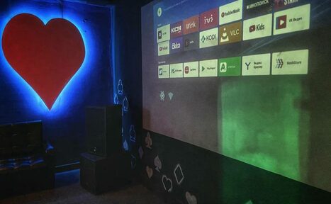 Арендовать Rave(Рейв). Комната с затемнённым освещением, чёрные стены с рисунками в виде сердечек и платиновых карт, большой экран с проекцией интерфейса приложений, красное сердце на стене с голубой подсветкой, кресло, аудиоколонка.