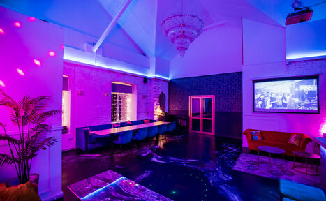 Арендовать Уютный лофт с отдельным входом и внутренним двором с барбекю. Большое помещение с высокими потолками, белыми кирпичными стенами и декоративными элементами. Мебель включает длинный стол с синими креслами и оранжевый диван. Освещение неоновое, синие и розовые тона. На стене висит монитор с изображением.
