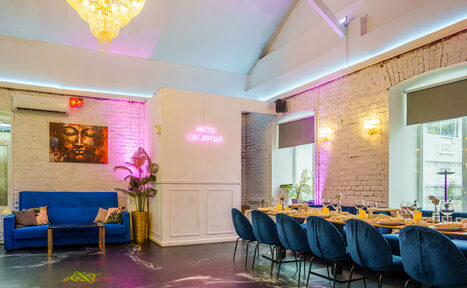 Арендовать Уютный лофт с отдельным входом и внутренним двором с барбекю. Просторное помещение с кирпичными стенами, большим столом и синими стульями. Сбоку диван с подушками. На стене неоновая надпись и картина. Потолок высокий, с люстрой, освещение разноцветное. На столе сервировка для обеда.