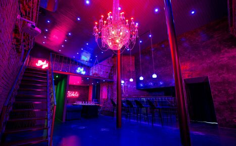 Арендовать Двухэтажный лофт с отдельным входом. Интерьер помещения с лестницей и барной стойкой. Красные и синие неоновые огни, хрустальная люстра, кирпичные стены. Подвесные светильники над барной стойкой с высокими стульями. Металлические перила и бетонные элементы. Имеются яркие неоновые надписи.