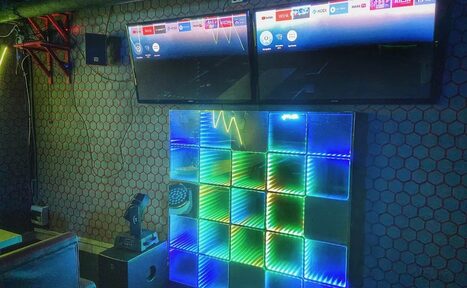 Арендовать Bee-lounge(Пчела-лаундж). Развлекательное пространство с двумя экранами на стене с сотами. Ниже экрана светящаяся панель с разноцветной подсветкой. Освещение неяркое, электрическое оборудование на стенах и полу. Цвета: синий, зеленый, желтый.