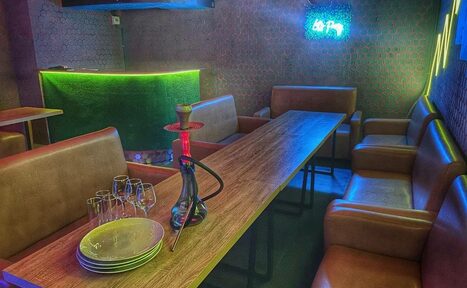 Арендовать Bee-lounge(Пчела-лаундж). Кафе-бар с угловым диваном и креслами из коричневой кожи вокруг длинного деревянного стола. На столе кальян и посуда. На стене светящаяся неоновая надпись. Стойка с зелёной подсветкой. Тёмные стены с узором.