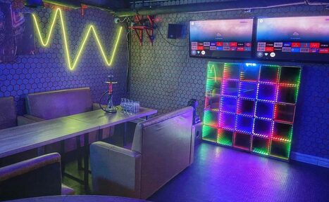 Арендовать Bee-lounge(Пчела-лаундж). Пространство для отдыха с длинным деревянным столом и мягкими диванами. На стене неоновая подсветка, рядом сдиванами кальян и стаканы. На другой стене два экрана и разноцветная светодиодная панель. Стены с ажурным узором.
