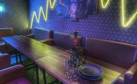 Арендовать Bee-lounge(Пчела-лаундж). Комната с длинным обеденным столом и коричневыми диванами вдоль стены. На столе кальян, тарелки и бокалы. Стена декорирована геометрическими обоями и неоновым освещением в виде диаграммы. На стене несколько картин.