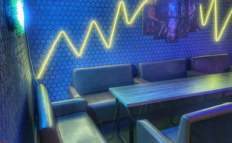 Арендовать Bee-lounge(Пчела-лаундж). Современное помещение с темной мебелью: два дивана и стол. На стенах плитка сине-черного цвета с узором "соты", неоновая подсветка в виде зигзага. На стене подвешены картины. Освещение мягкое, интерьер в хорошем состоянии.