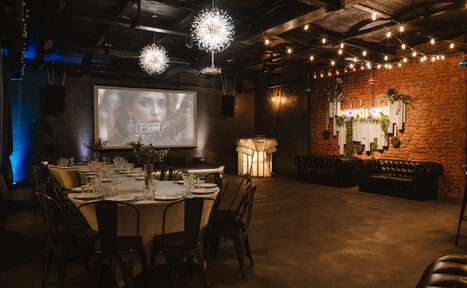 Арендовать Nord Loft(Норд Лофт). Пространство с кирпичной стеной, деревянным потолком и бетонным полом. В центре круглый стол с черными стульями, сервирован стеклянной посудой. На стене экран с изображением, рядом кожаные диваны и неоновая вывеска. Потолочные светильники и гирлянды создают мягкое освещение.