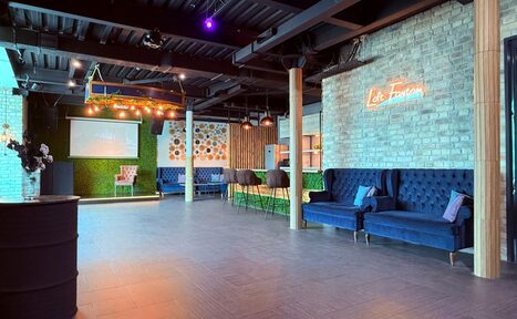 Арендовать Лофт Фантом. Многофункциональное пространство с открытой планировкой, кирпичными стенами, синими диванами, барными стульями. На стенах неоновая вывеска и зеленая зона. Потолок металлический, освещение подвесное. На полу темное деревянное покрытие.