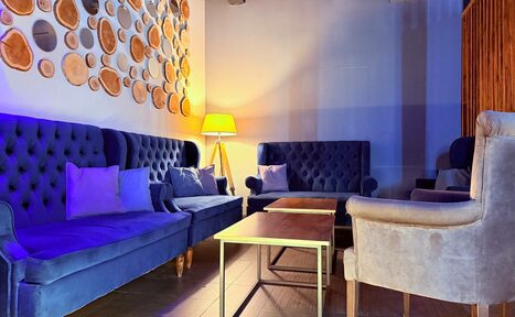 Арендовать Лофт Фантом. Простор с синими диванами и серыми креслами, деревянные журнальные столики, лампа с желтым абажуром, стена украшена круглыми деревянными элементами, пол из светлого дерева, мягкое освещение.