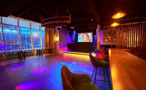 Арендовать Лофт Фантом. Просторное помещение с большими окнами и видом на город. Стены из кирпича, на одной из них экран с изображением. Потолок темный с декоративными элементами. Комфортные стулья у деревянной барной стойки. Уютное цветное освещение.