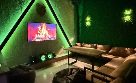 Арендовать Лофт Фантом. Лаунж-зона с мягкими диванами, деревянным столом, двумя креслами-грушами. На стене телевизор с изображением камина, стильное зелёное освещение. Стена с растениями, акустическая система. Настенная фактура и текстуры под дерево.
